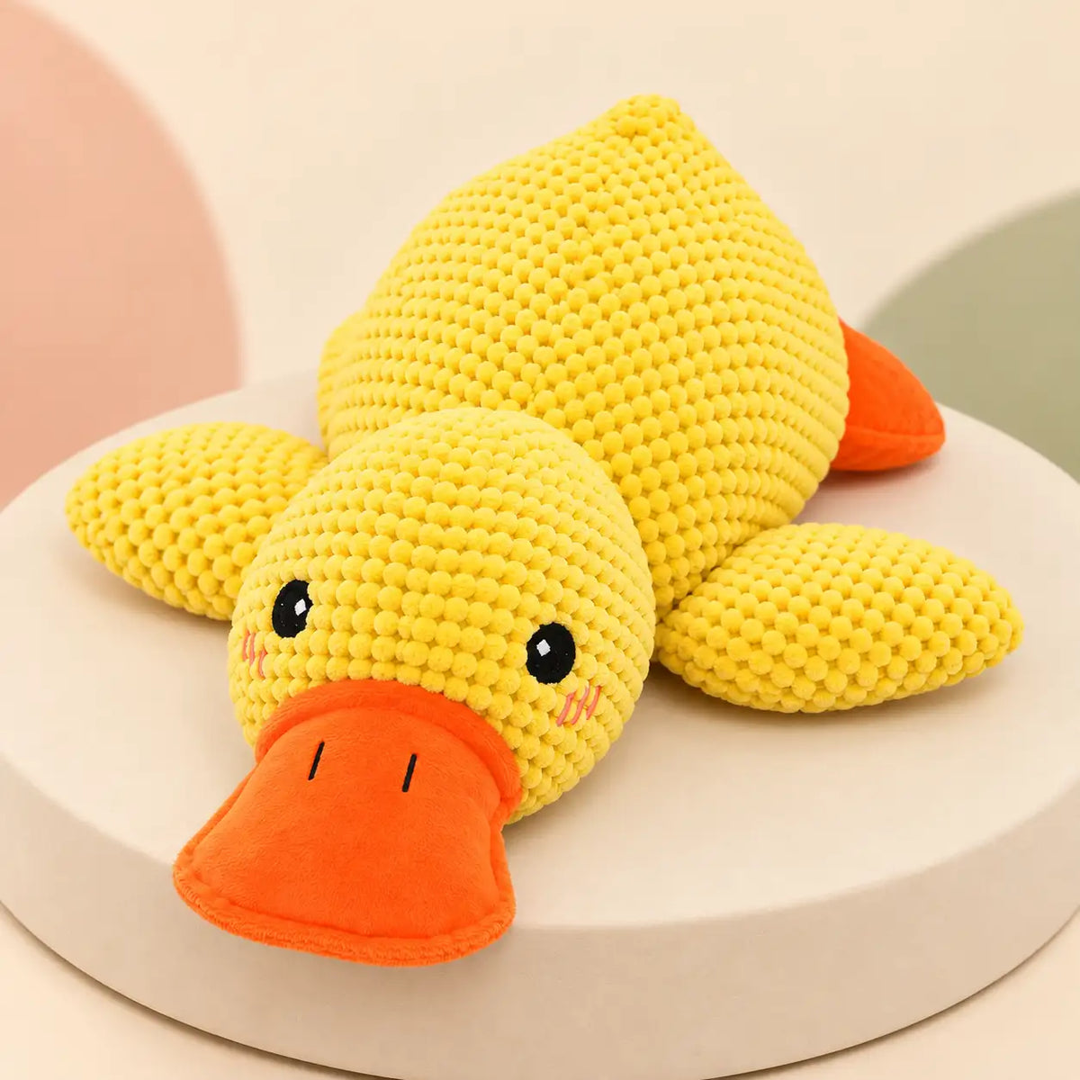 A yellow crocheted duck plush toy with an orange beak and orange feet, resting on a round white pedestal against a soft pastel background.