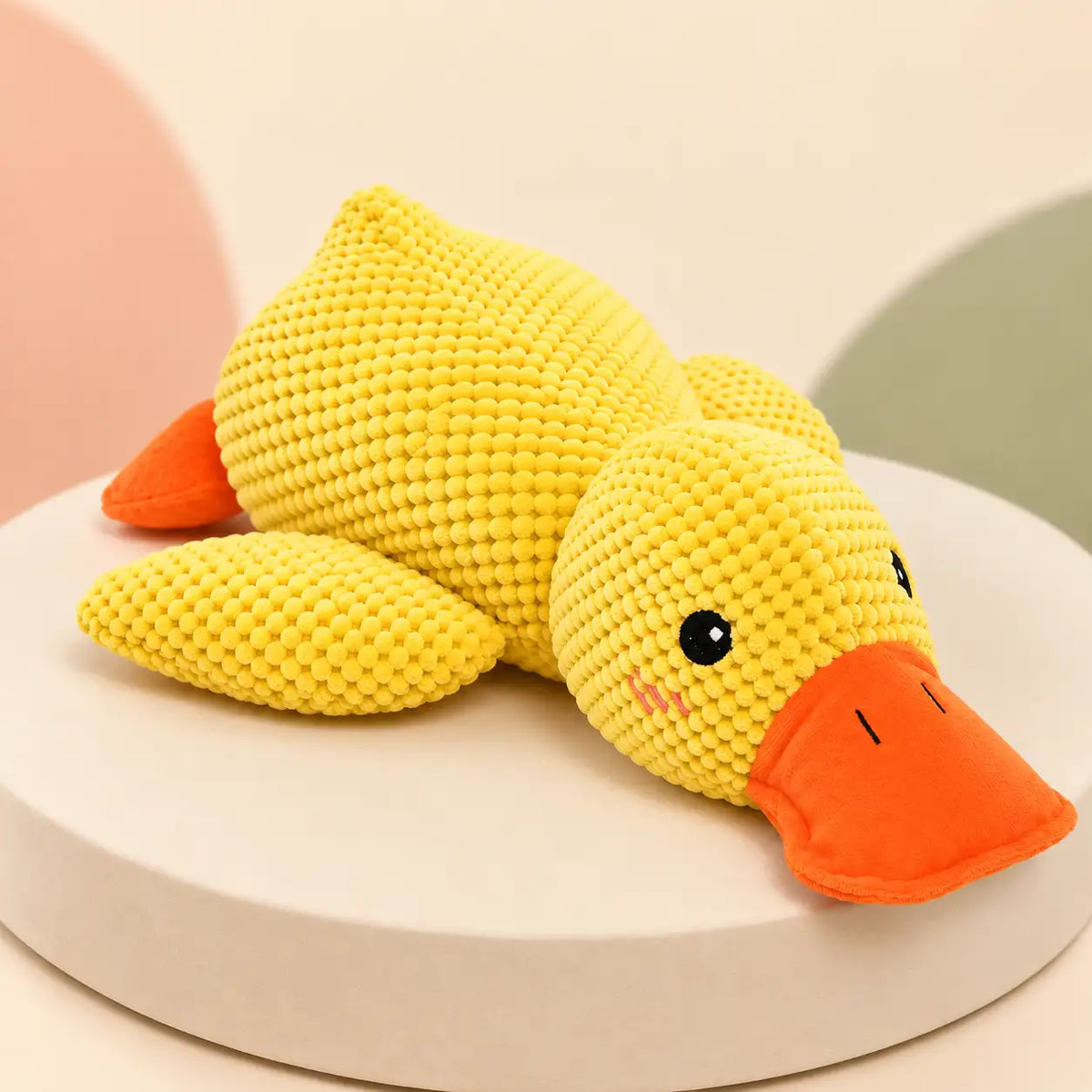 A yellow crocheted duck toy with an orange beak and orange tail, resting on a round white pedestal against a soft pastel background.