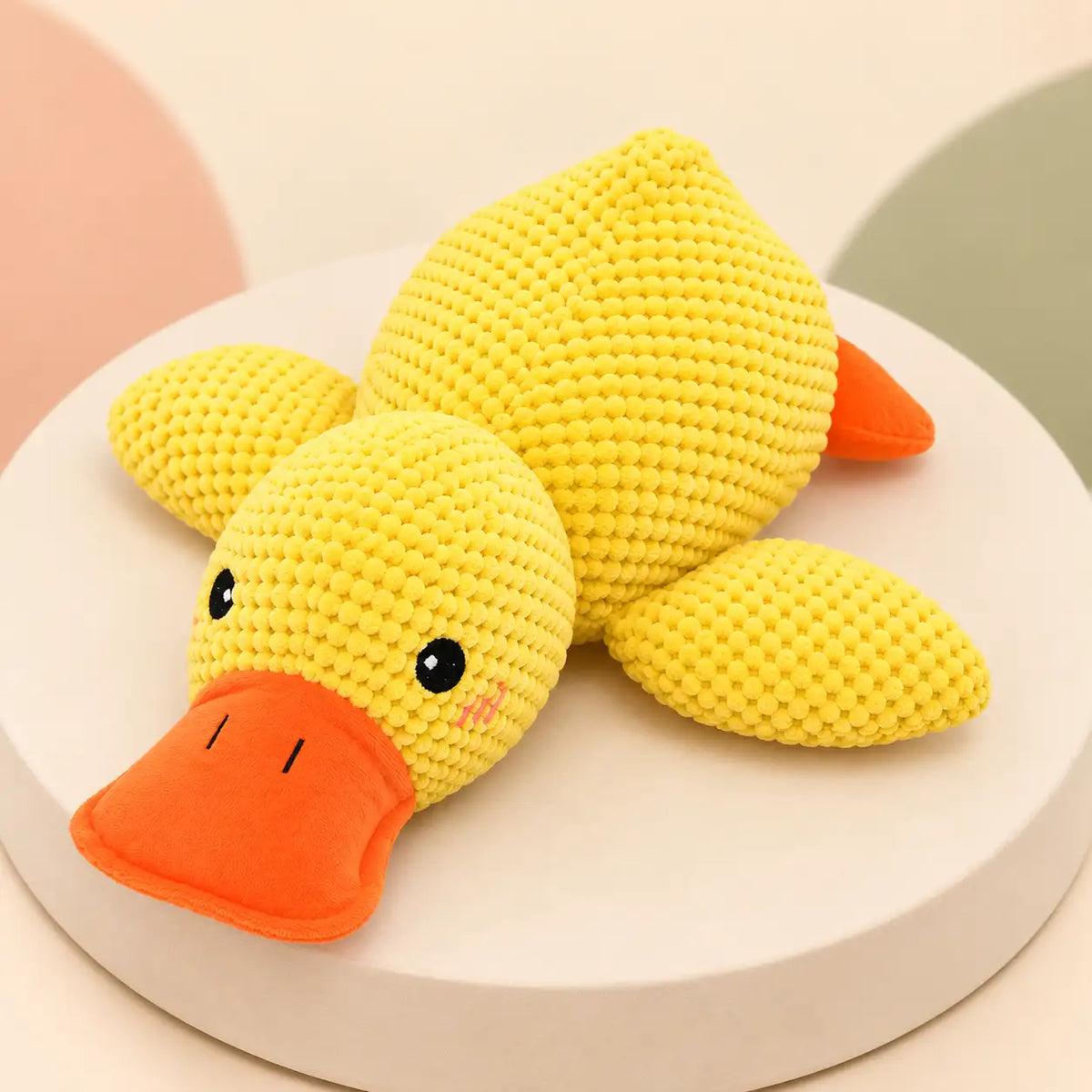 A yellow crocheted duck toy with an orange beak and orange tail fin, resting on a round white pedestal against a soft pastel background.