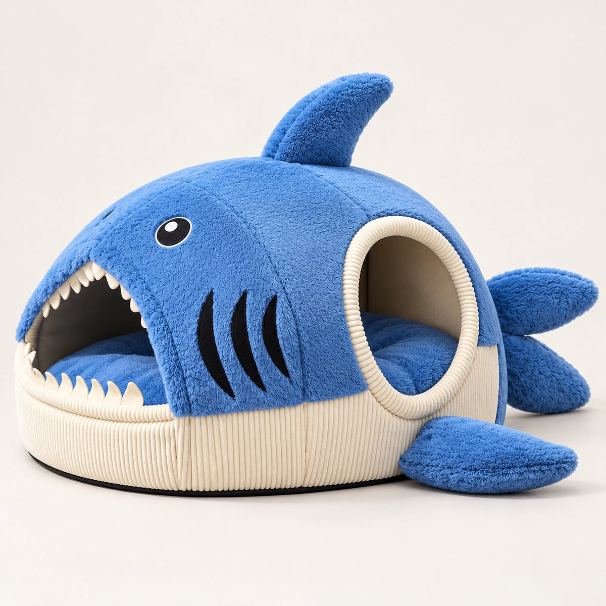 A plush shark-shaped pet bed in blue and cream, featuring a circular opening with a ribbed cream border, a shark face with black eyes and teeth details, and three blue pectoral fins on the front.
