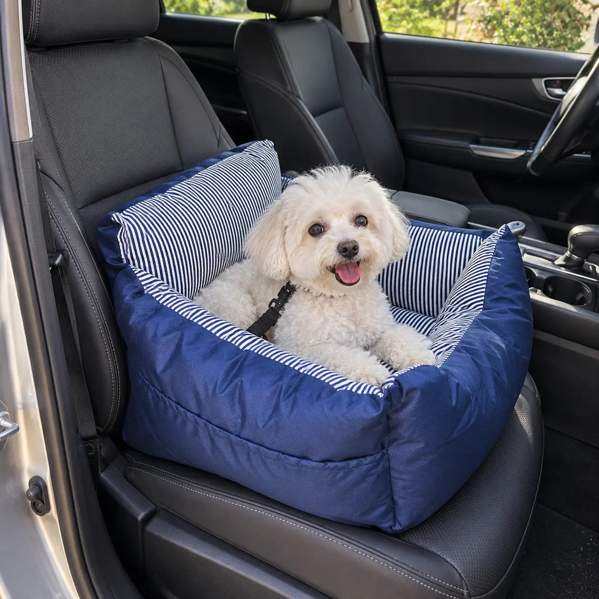 A small white dog sits inside a blue car seat pet bed with striped fabric lining, placed on a car's front seat.